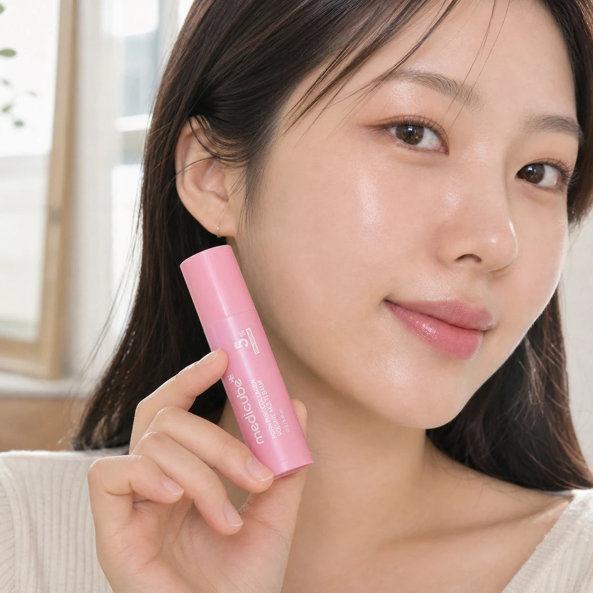Woman holding medicube PDRN Pink Collagen Volume Multi Balm 10g beside her cheek