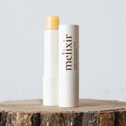 ProtoClinical k-beauty melixir Vegan Lip Butter 3.9g #01 Agave(Clear) open clear lip butter stick beside a capped white tube on a cut wood surface against a pale gray background
