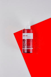 ProtoClinical k-beauty PAUL MEDISON Signature Body Mist 211ml #Baby Powder single translucent pink-red body mist bottle standing upright against an angled red panel with water droplets on a white background