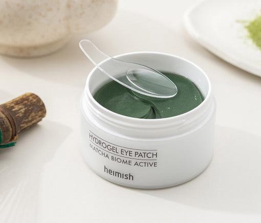 ProtoClinical k-beauty heimish Matcha Biome Hydrogel Eye Patch 1.4g X 60pcs open white round jar filled with dark green crescent eye patches on a pale green water-ripple background