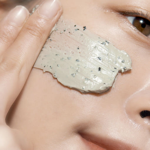 ProtoClinical k-beauty [Pyunkang Yul] Calming Pore Clear Wash Off Pack 100g close-up of a gray clay mask spread across a cheek with visible fingertips and dark exfoliating granules