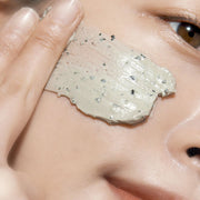 ProtoClinical k-beauty [Pyunkang Yul] Calming Pore Clear Wash Off Pack 100g close-up of a gray clay mask spread across a cheek with visible fingertips and dark exfoliating granules