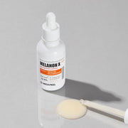 ProtoClinical k-beauty MEDIPEEL Melanon X Ampoule Light 30ml pale beige ampoule packaging with a dark gray oval detail on a light background