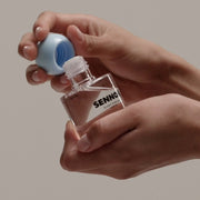 ProtoClinical k-beauty SENNOK Glossy Everyday Hair Oil 60ml shown in a close-up with two hands holding the clear bottle at an angle and removing the round blue cap on a soft beige background