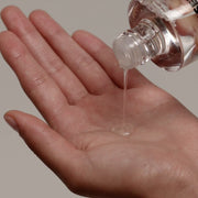ProtoClinical k-beauty SENNOK Glossy Everyday Hair Oil 60ml in a close-up of the open clear bottle pouring translucent oil onto an open palm against a soft beige background