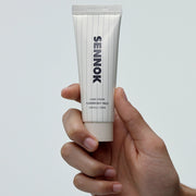 ProtoClinical k-beauty SENNOK Hand Cream Flower But Wild 50ml shown upright in a hand, with a white ribbed tube and beige cap against a pale blue background