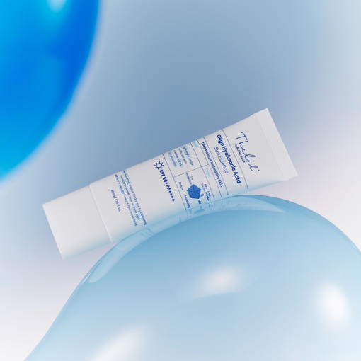 ProtoClinical k-beauty [THE LAB by BLANC DOUX] Oligo Hyaluronic Acid Sun Essence SPF50+ PA++++ 40ml white tube lying diagonally across pale blue balloons on a soft light background