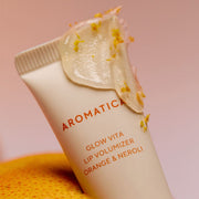ProtoClinical k-beauty AROMATICA Glow Vita Lip Volumizer Orange & Neroli 10g close-up of a beige squeeze tube topped with glossy lip gel and orange peel on a soft pink background