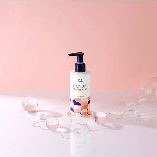 ProtoClinical k-beauty Foellie Capsule Feminine Wash 200ml upright bottle on a glossy white surface with translucent spheres and spilled liquid against a pink background