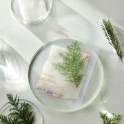 ProtoClinical k-beauty mixsoon Daily Mask Pack Wednesday 20ml X 10ea clear sheet mask pouch resting on a round glass plate with green cypress sprigs and clear dishware on a bright white surface