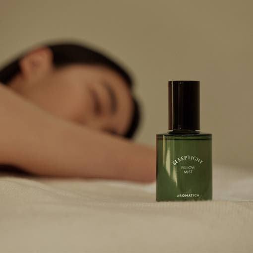 ProtoClinical k-beauty AROMATICA Sleep Tight Pillow Mist 100ml dark spray bottle beside a cream pillow in a warm brown bedroom scene