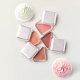 ProtoClinical k-beauty CLIO Essential Lipcheek Tap Blur 4.5g (4colors) flat lay of four triangular balm compacts in pink and peach tones beside two cupcakes on a pale background