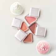 ProtoClinical k-beauty CLIO Essential Lipcheek Tap Blur 4.5g (4colors) flat lay of four triangular balm compacts in pink and peach tones beside two cupcakes on a pale background