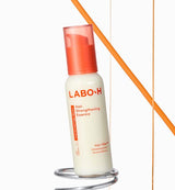 ProtoClinical k-beauty LABO-H Hair Strengthening Essence 75ml as an ivory essence bottle with a translucent coral cap leaning on a chrome ring against a white background with an orange diagonal line