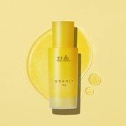 ProtoClinical k-beauty HANYUL Moonlight Yuja C Serum 30ml upright yellow serum bottle over a glossy yellow serum pool with two small droplets on a pale yellow background